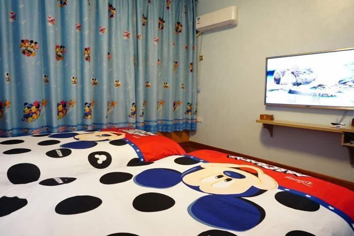 A hotel room with a Mickey Mouse themed bedspread, a television on the wall, and a curtain with cartoon characters.