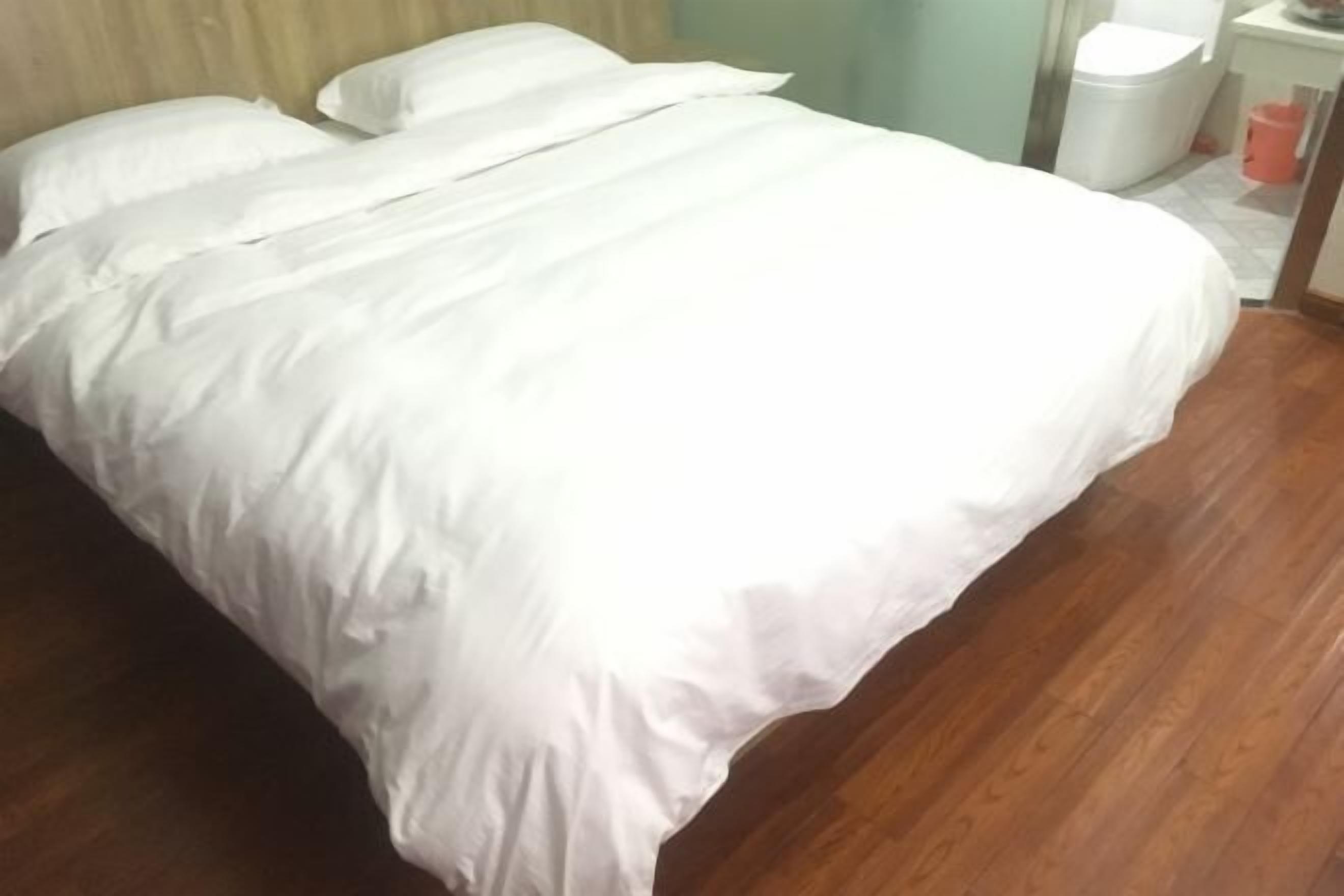 A neatly made bed with white bedding in a room with wooden flooring and a white trash bin.