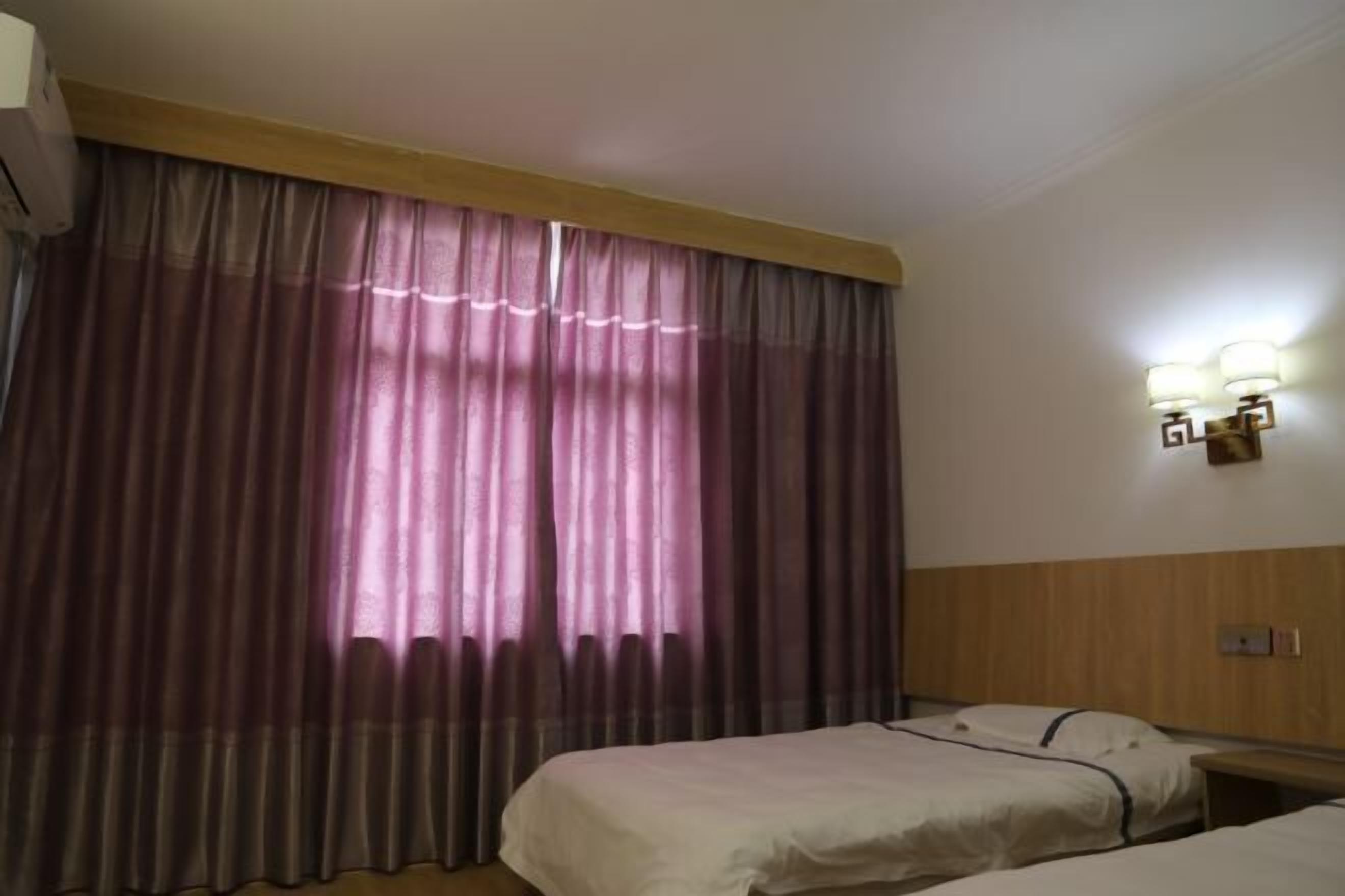 Hotellihuone, jossa on kaksi sänkyä, suuri ikkuna, jossa on verhot, seinään kiinnitetty valaisin ja puinen sängynpääty.