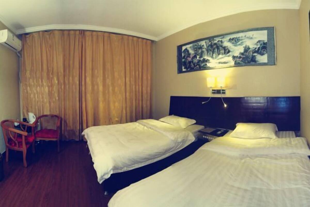A hotel room with two beds, a chair, a painting on the wall, and an air conditioning unit.