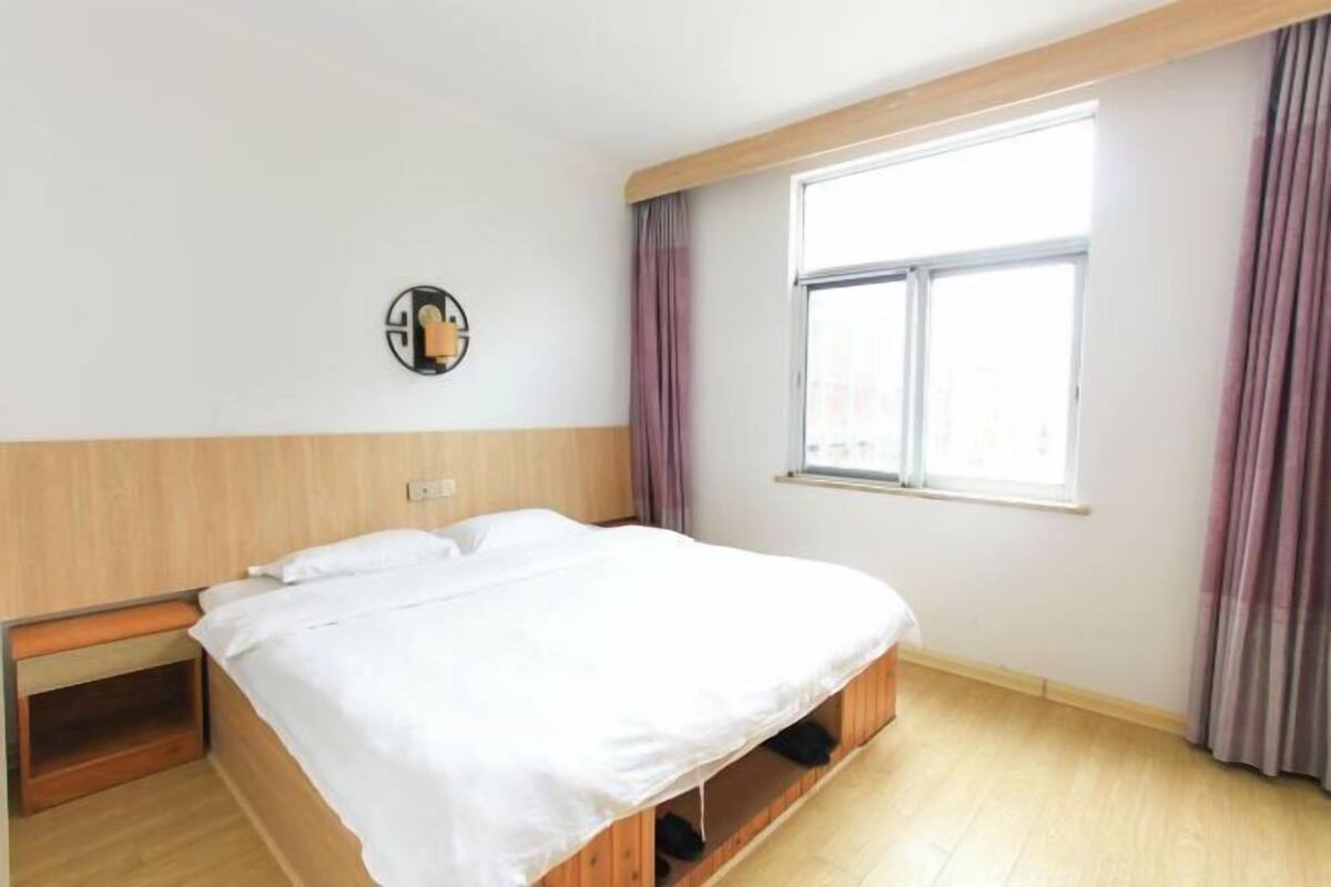 A single bed with a wooden headboard, a window with curtains, and a wall-mounted light fixture.
