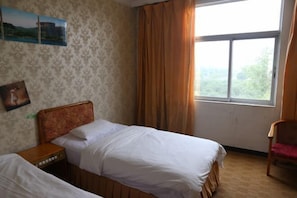 A hotel room with two beds, a wooden headboard, a nightstand, a chair, and a window with curtains. - ChunHui Inn Hotel (Wuyishan)