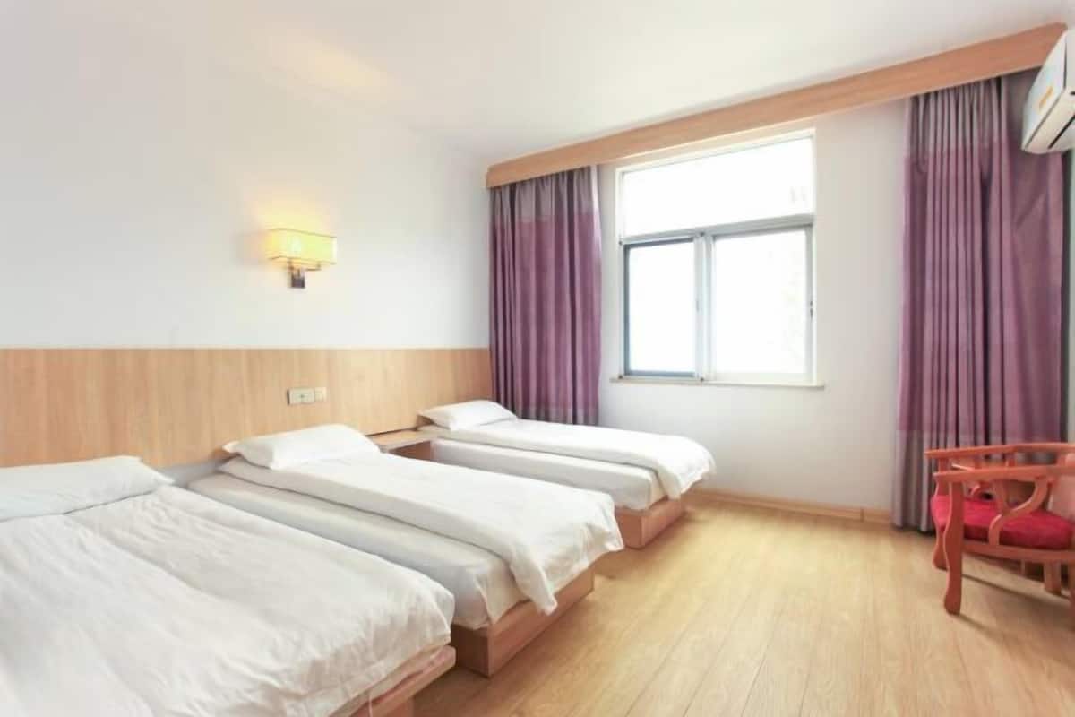 A hotel room with three beds, a wooden headboard, a window with curtains, a wall-mounted lamp, and a wooden chair.