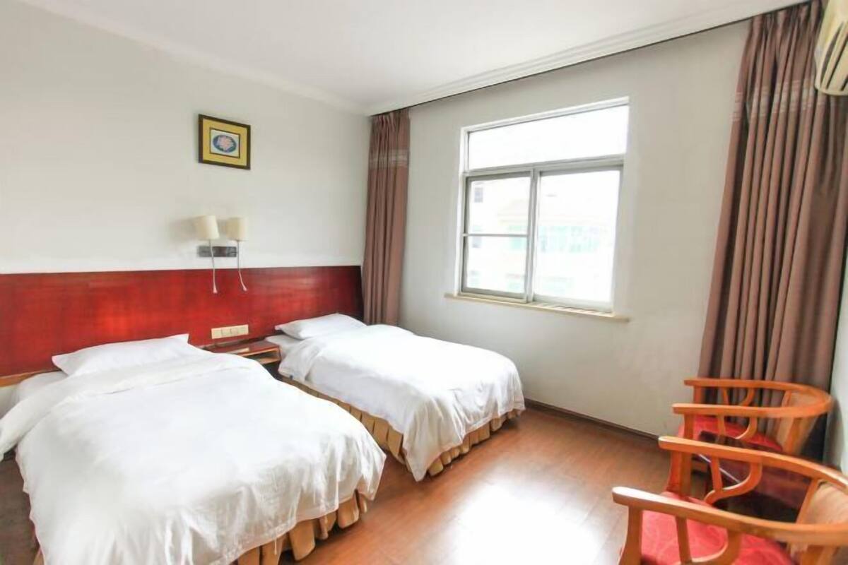 A hotel room with two beds, a window with curtains, a wall-mounted lamp, and a wooden chair.