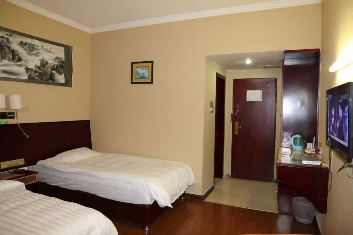 A hotel room with two beds, a wooden headboard, a painting on the wall, a small framed picture, a desk with a computer, and a door leading to another room.