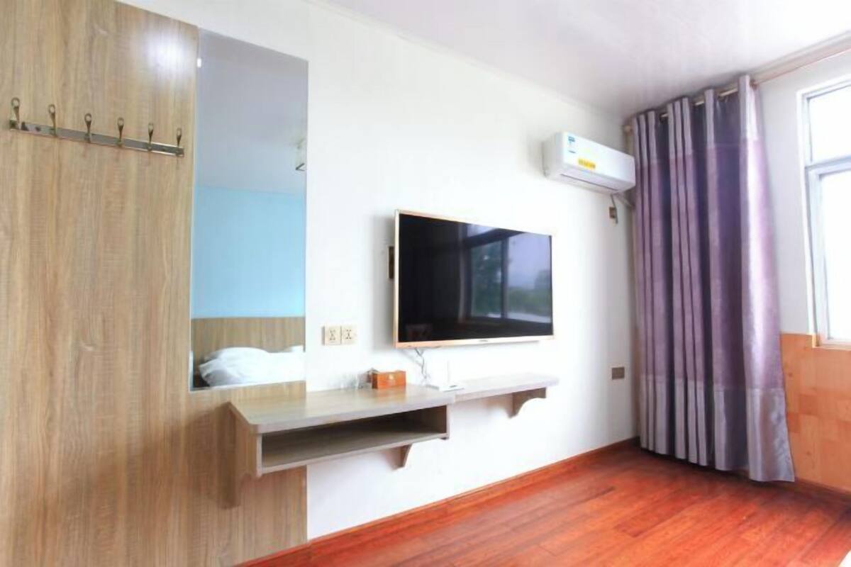 A room with a flat-screen TV mounted on the wall, a wooden wardrobe, a built-in shelf, and a window with curtains.