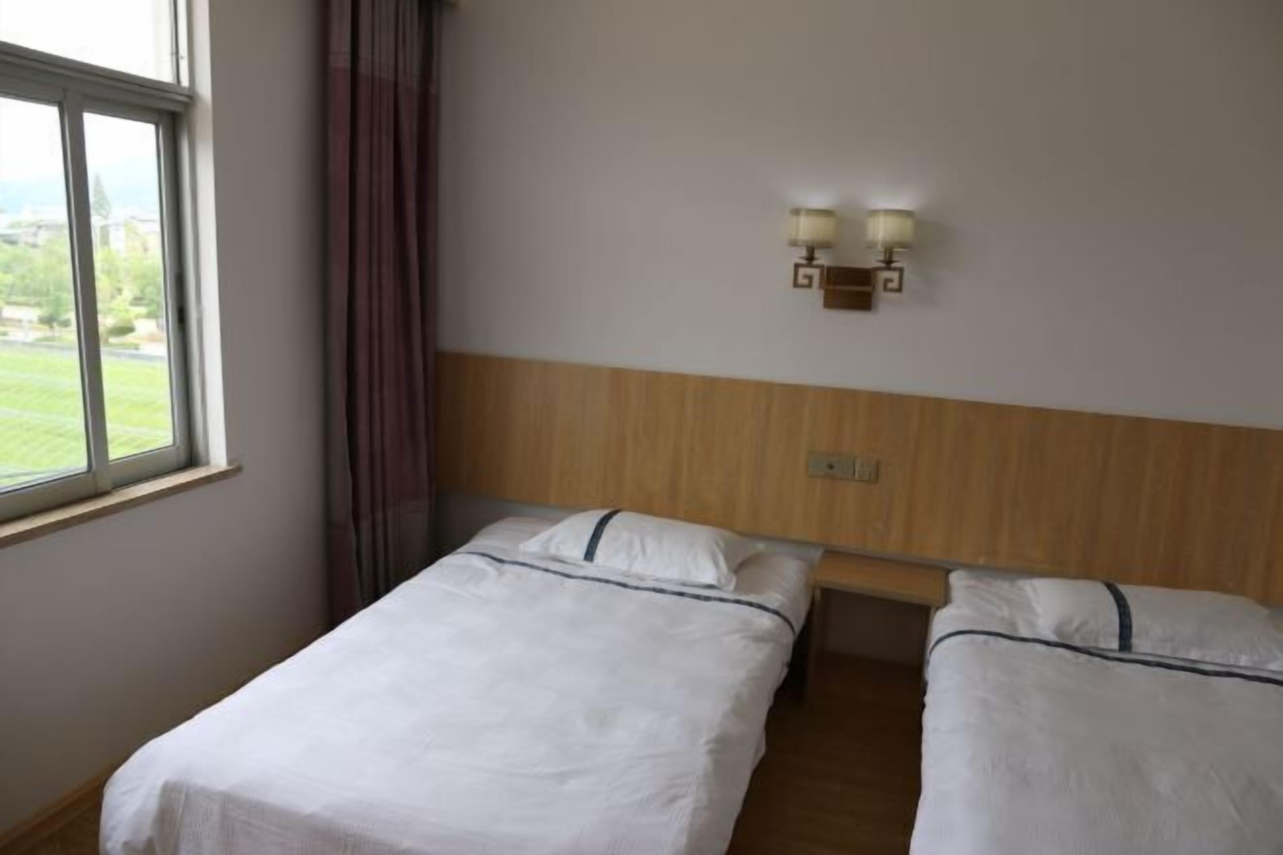 A hotel room with two single beds, a window with curtains, and a wall-mounted light fixture.