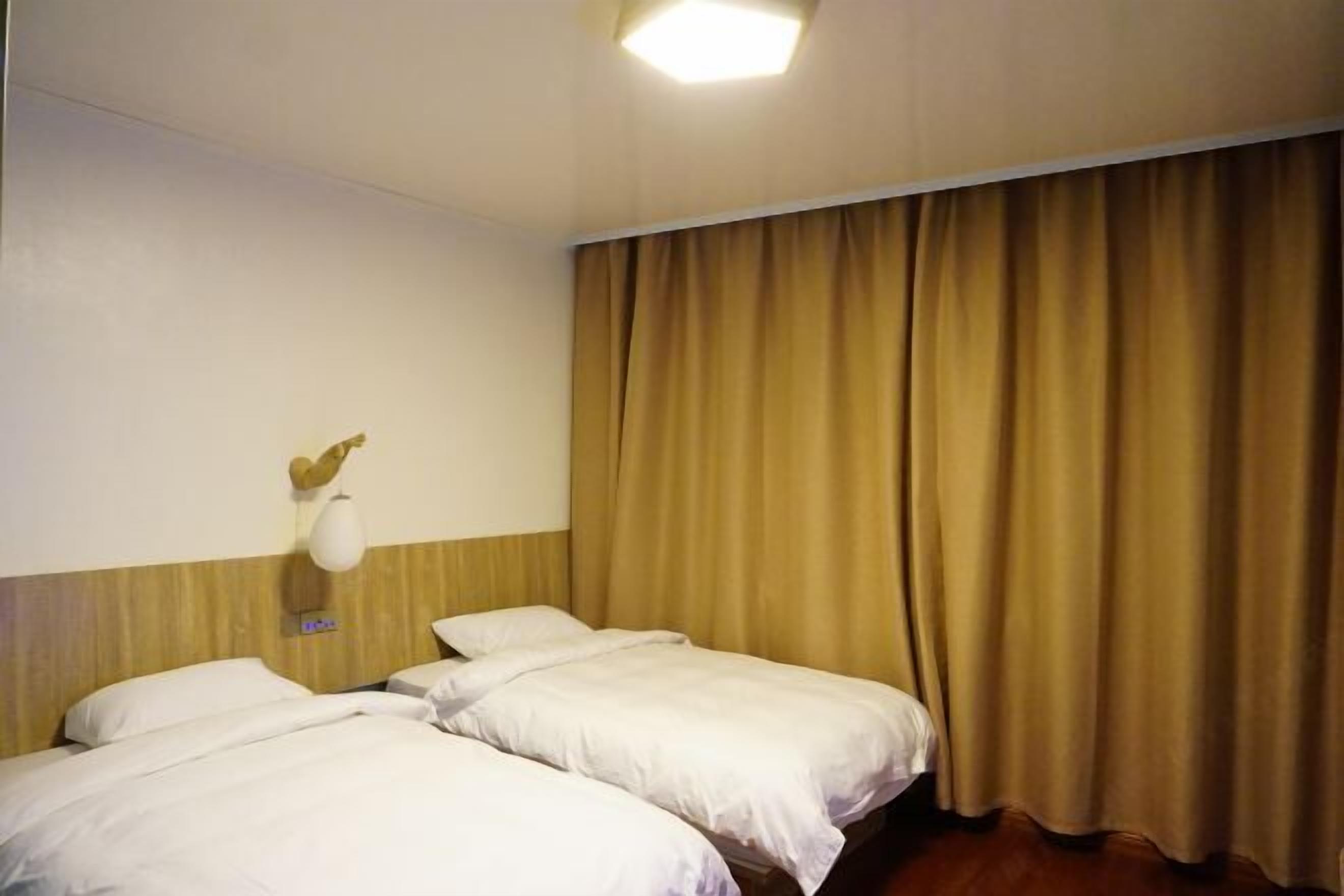 A hotel room with two beds, a wooden headboard, a wall-mounted light fixture, and a large window with curtains.