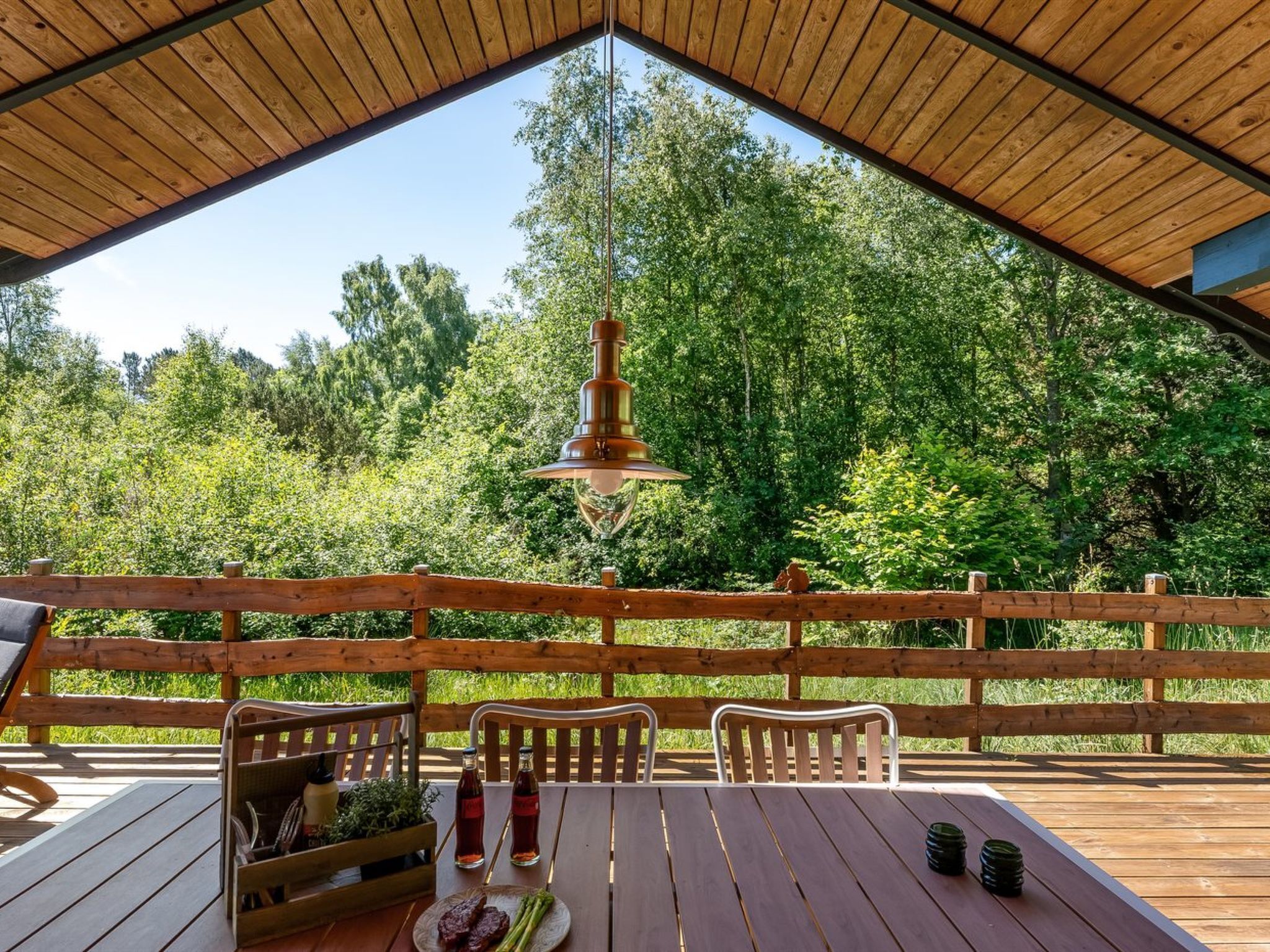 Une terrasse en bois avec une table dressée pour le repas, un luminaire suspendu et une vue sur une zone boisée.