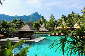 A resort pool area with lounge chairs, umbrellas, and thatched-roof huts, surrounded by lush greenery and palm trees. - DLF Stays (Gurugram)