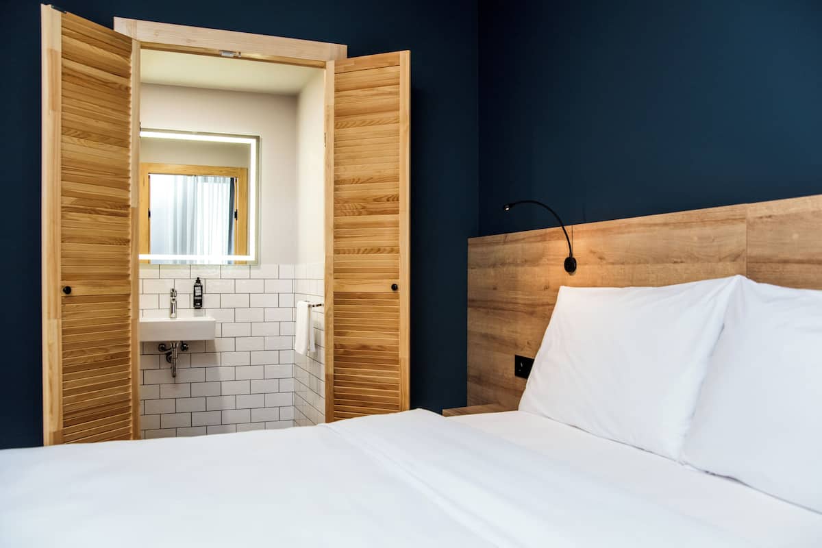 Une chambre d’hôtel avec une tête de lit en bois, un lit blanc, une armoire en bois avec un miroir, et un lavabo blanc avec une serviette accrochée au mur.