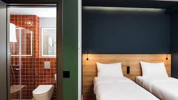 Quarto de hotel com cabeceira de madeira, duas camas, chuveiro e vaso sanitário.