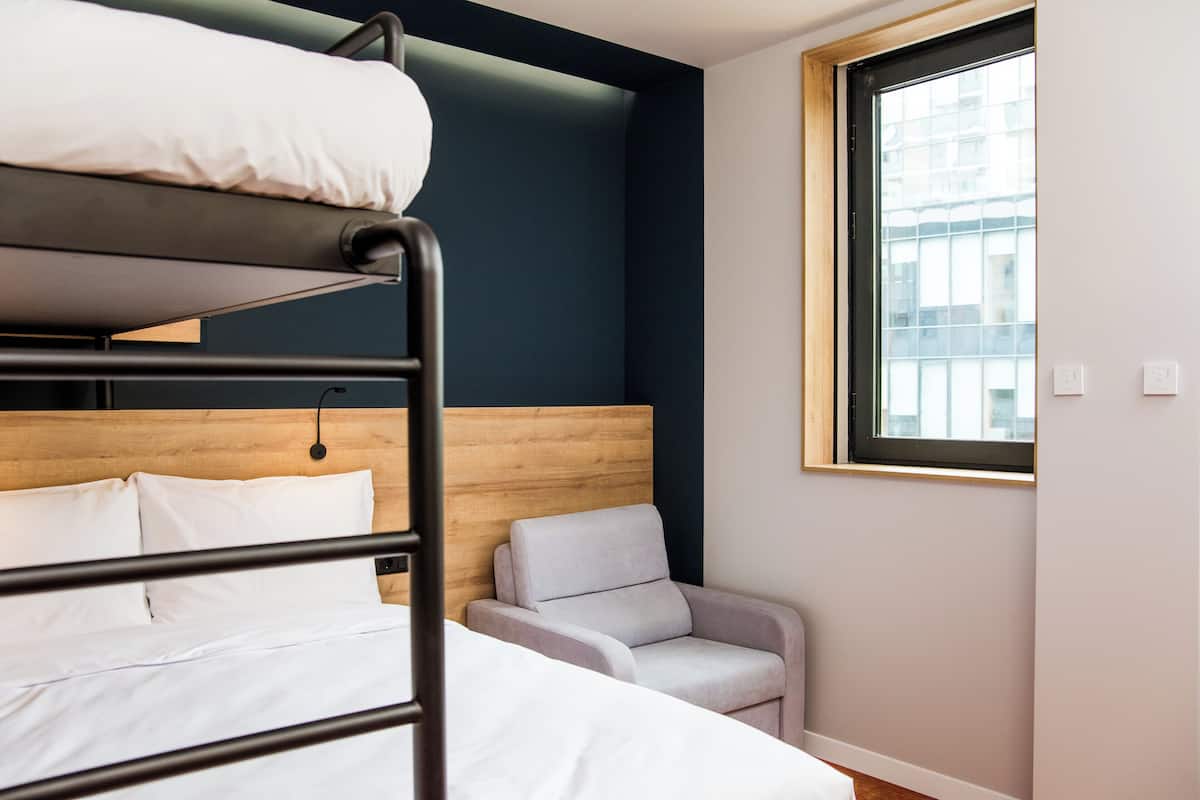 Un lit superposé avec une literie blanche, une tête de lit en bois, un petit fauteuil avec un coussin et une fenêtre donnant sur des immeubles.