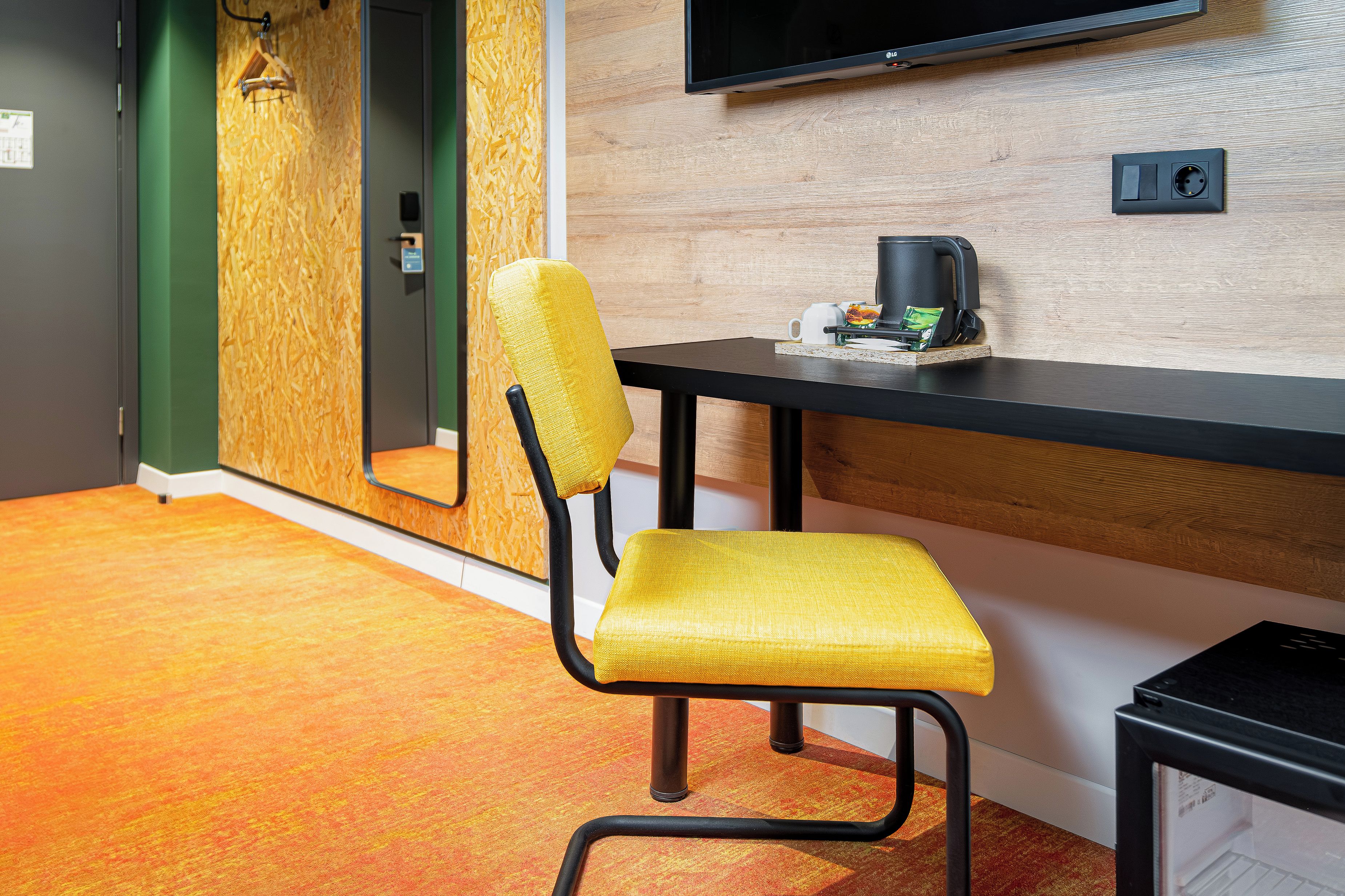 A modern hotel room with a yellow chair, a black desk, a television mounted on the wall, and a small black appliance on the desk.