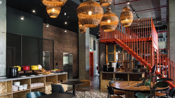 A modern restaurant with a brick wall, wooden tables, wicker pendant lights, and a red staircase.