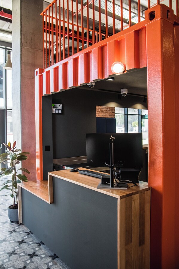 A reception desk with a computer, a potted plant, and a view of the outdoors through a window.