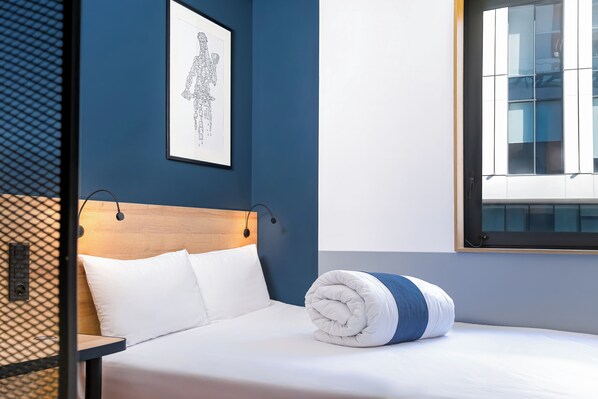 Una cama bien hecha con ropa blanca y una toalla enrollada, un cabecero de madera, una obra de arte enmarcada en la pared y una ventana con vistas a edificios.