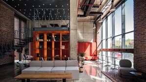 A modern lounge area with a sofa, wooden table, and bar stools. There are large windows, a staircase, and a brick wall.