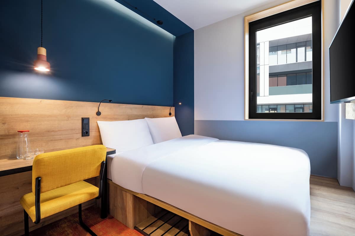 Une chambre d’hôtel moderne avec une tête de lit en bois, une chaise jaune, un lit blanc et une grande fenêtre donnant sur le bâtiment.