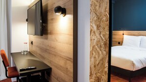 A hotel room with a wooden wall, a flat-screen TV mounted above a table with a remote control, and a bed with white bedding.
