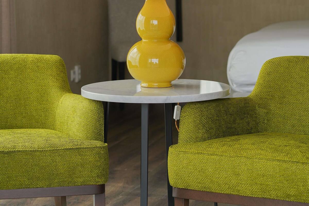 A hotel room with two green upholstered chairs, a small round table, and a yellow lamp.