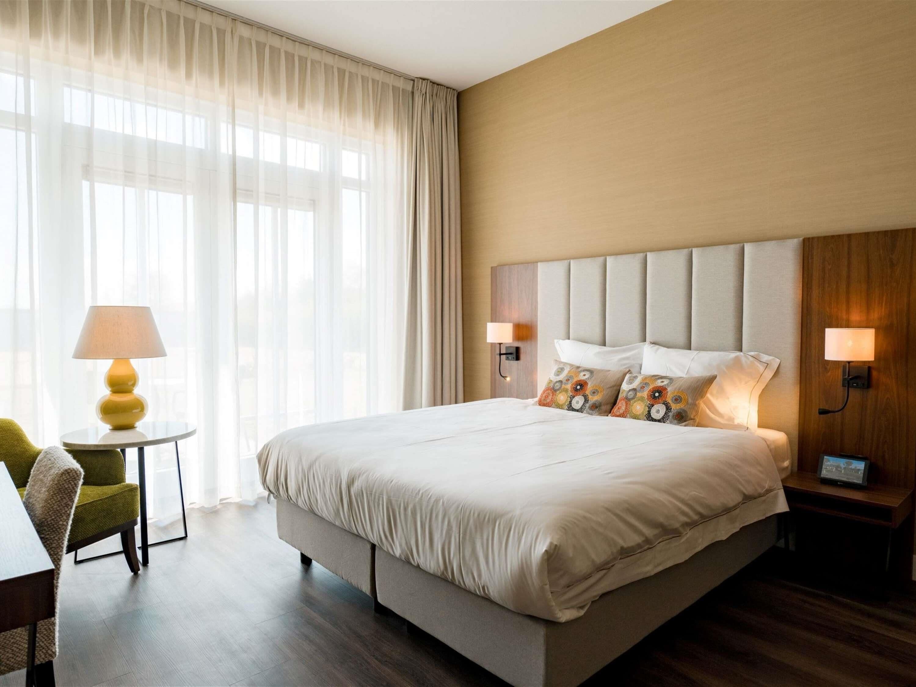 A hotel room with a large bed, two bedside lamps, a small table with a lamp, a chair, and a view through sheer curtains.