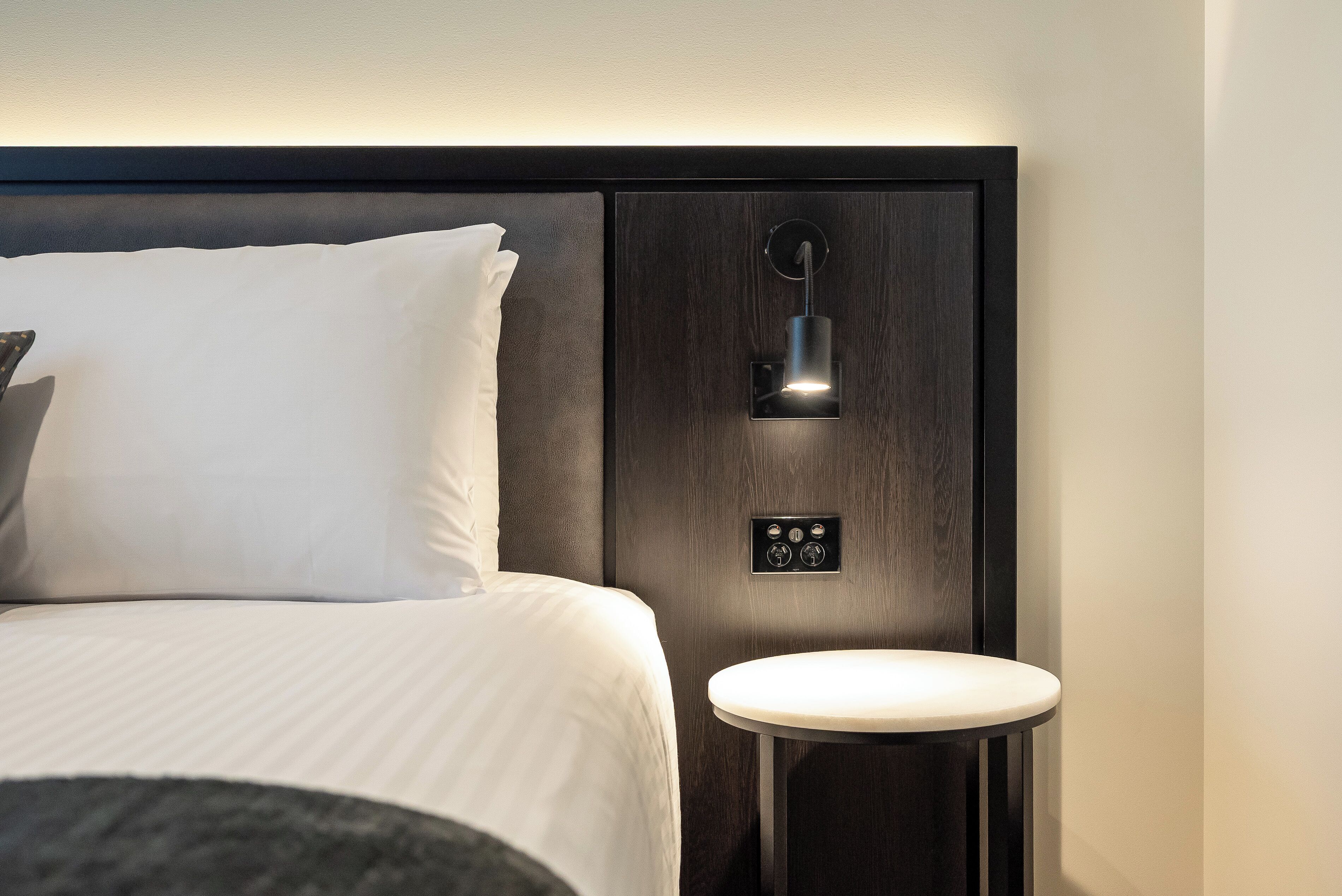A modern hotel room with a neatly made bed, a bedside table, and a wall-mounted light fixture.