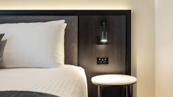 A modern hotel room with a neatly made bed, a bedside table, and a wall-mounted light fixture.