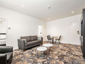 Quarto de hotel moderno com sofá cinza, duas mesas redondas, carpete estampado e porta branca.