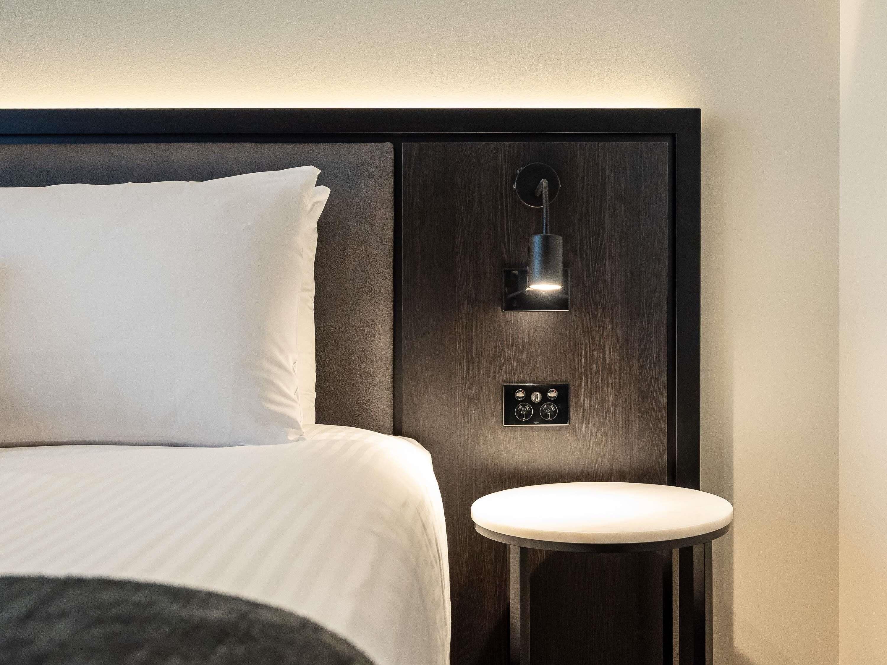 Una habitación de hotel con cama, mesita de noche, lámpara de pared y toma de corriente.