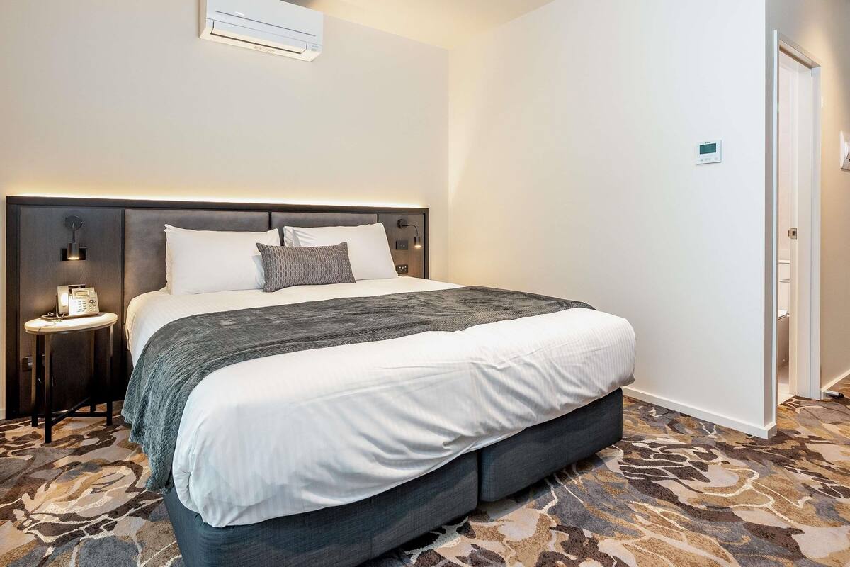 A hotel room with a large bed, bedside tables, a wall-mounted air conditioner, and a door.