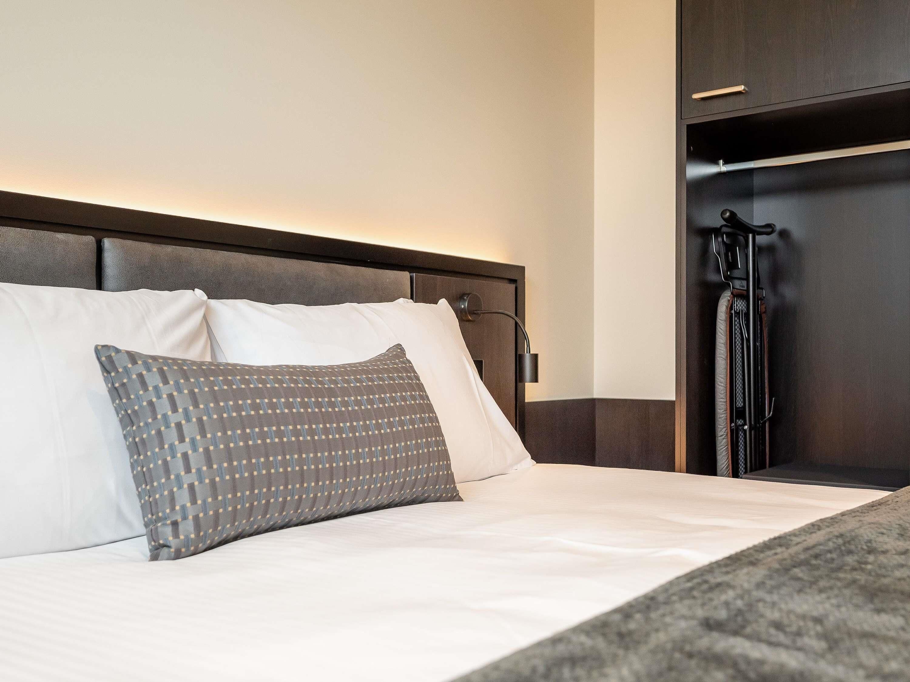 Una cama bien hecha con almohadas blancas y estampadas, un cabecero y un armario de madera oscura con perchero.