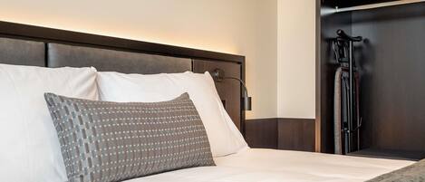 Una cama bien hecha con almohadas blancas y estampadas, un cabecero y un armario de madera oscura con perchero.