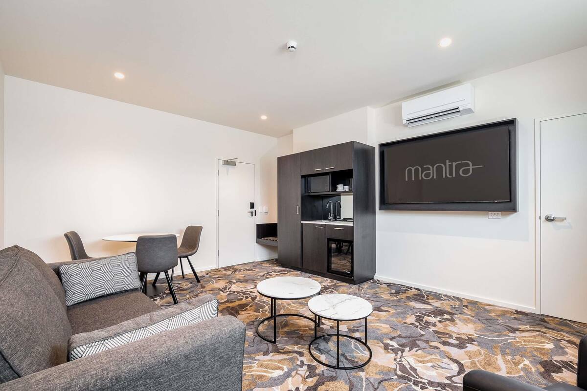 A modern living room with a sofa, two chairs, a small table, a kitchen area, and a mounted TV.
