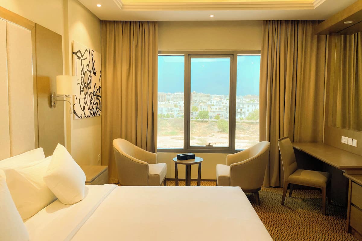 Une chambre d’hôtel avec un grand lit, deux fauteuils, un bureau et une vue sur le paysage urbain par la fenêtre.