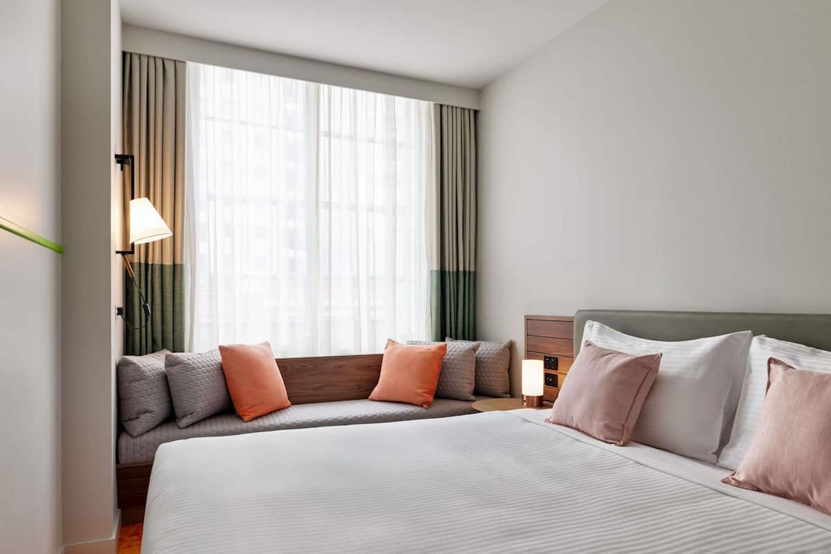 Habitación de hotel con una cama grande, un sofá cama, una lámpara y vistas de la ciudad a través de cortinas translúcidas.