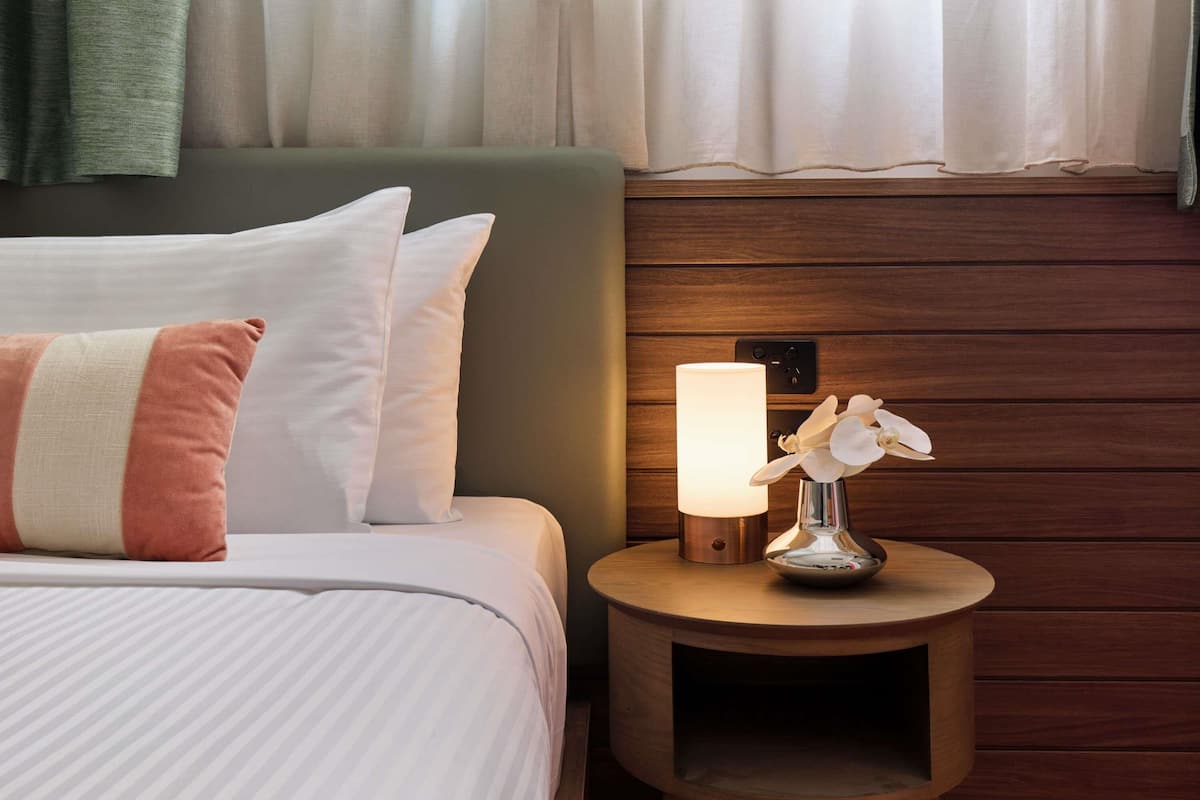 Una cama bien hecha con almohadas blancas y rosadas, una mesita de noche con una lámpara y orquídeas, y paneles de madera en la pared.