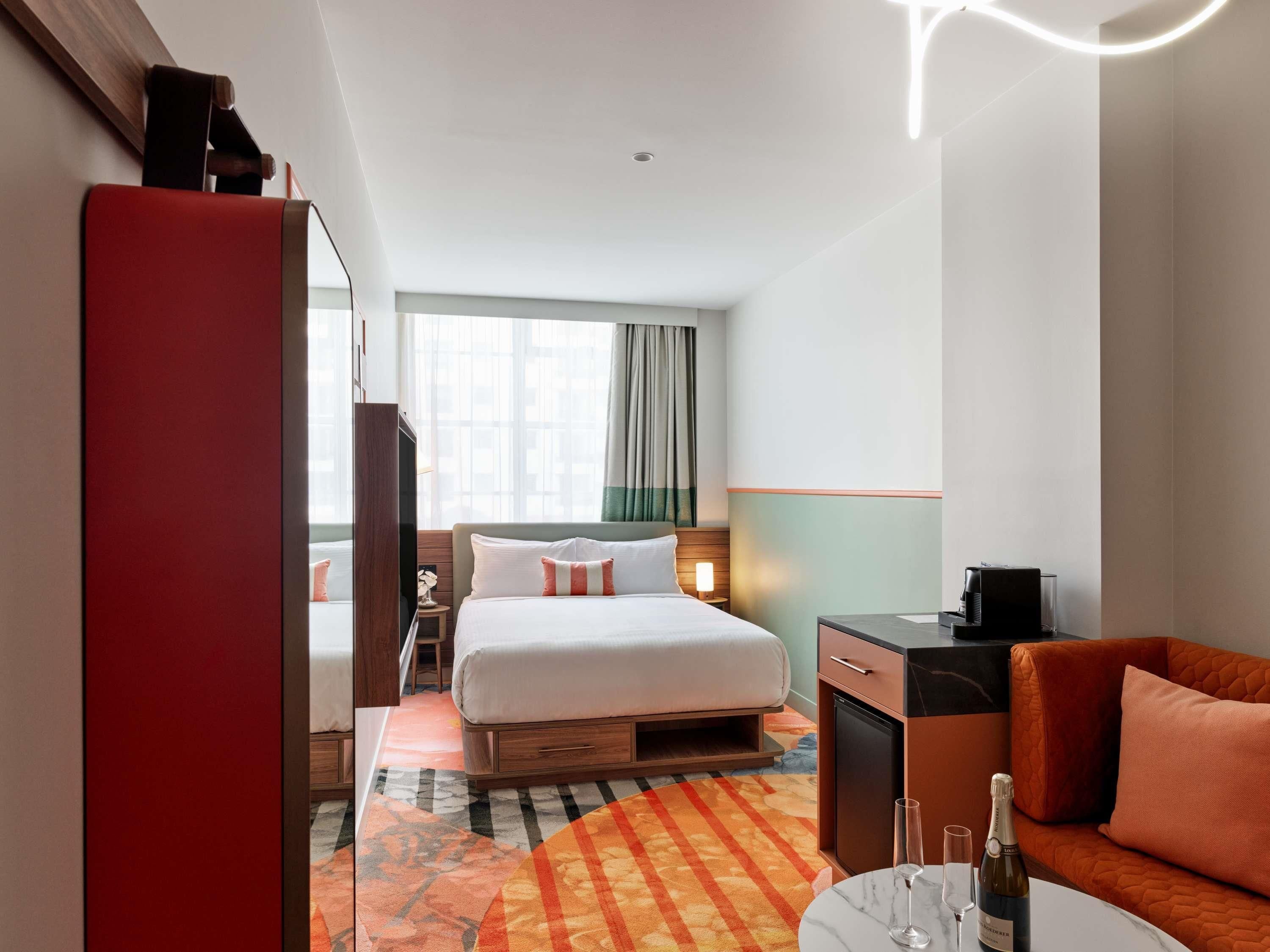 Camera d'albergo con un letto, una scrivania, una sedia, un tavolino con una bottiglia di champagne e calici, e un tappeto colorato.