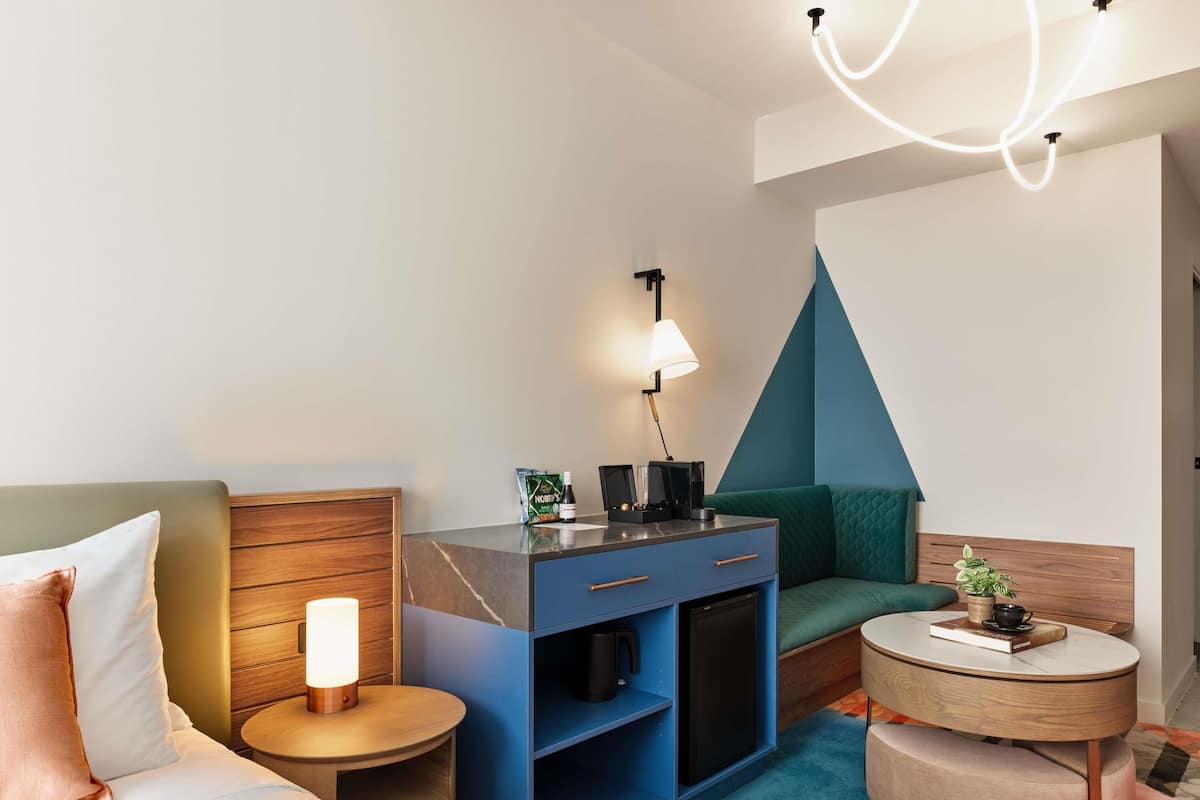 Una habitación de hotel moderna con una cama, un escritorio con lámpara, una mesita, un sofá y un diseño de pared geométrico único.