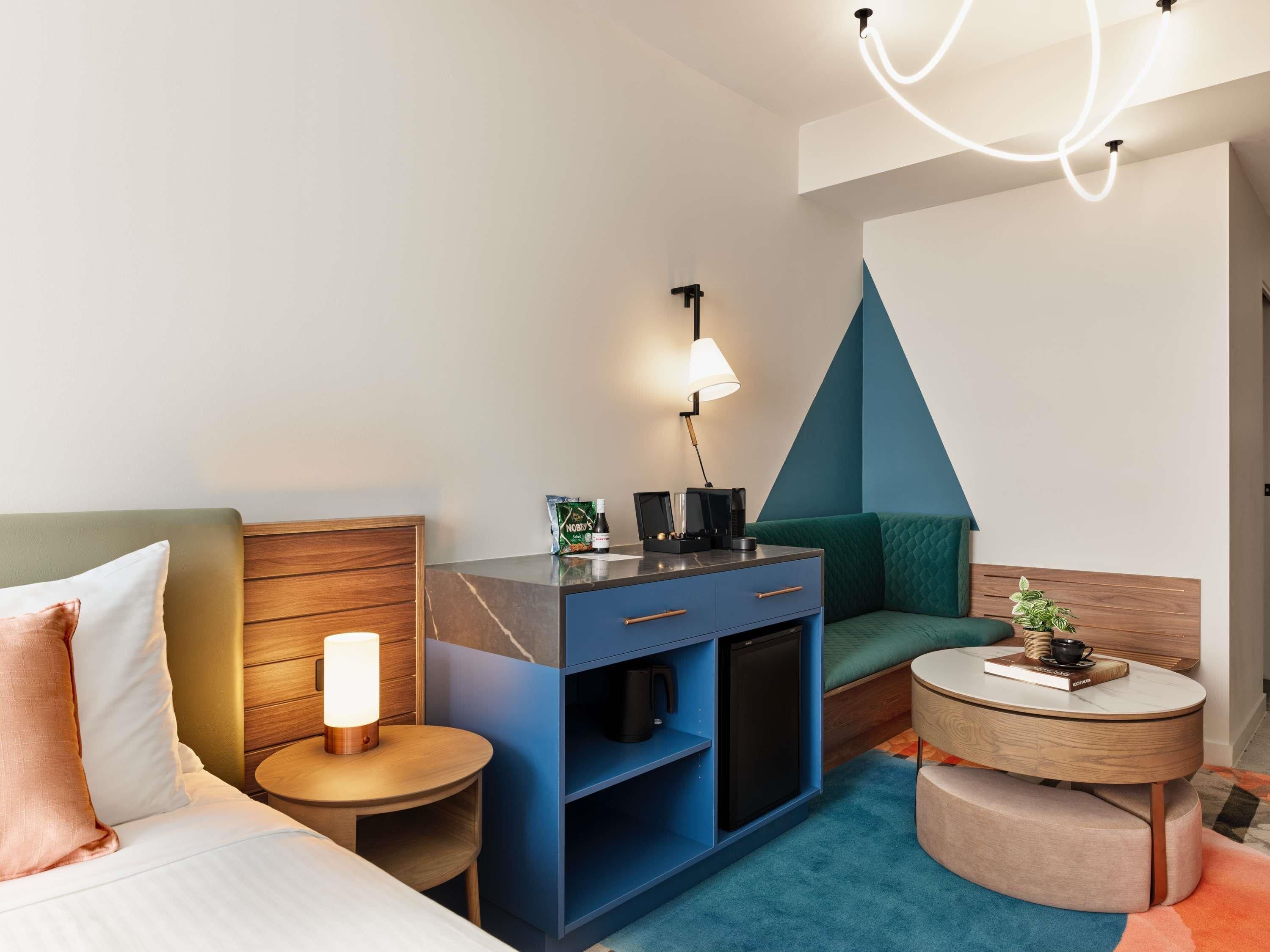 A modern hotel room with a bed, a desk with a lamp, a small table, a sofa, and a unique geometric wall design.