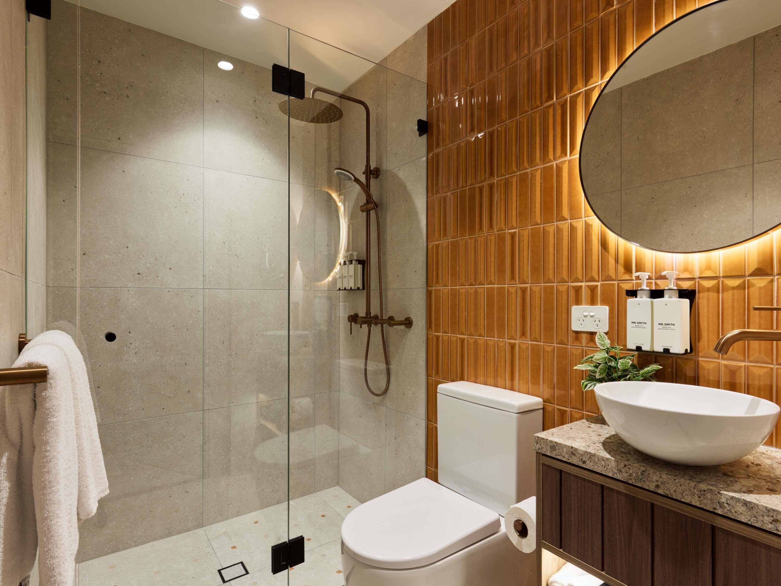 Un baño moderno con ducha de vidrio, inodoro, lavamanos de gran tamaño y espejo sobre el lavamanos.