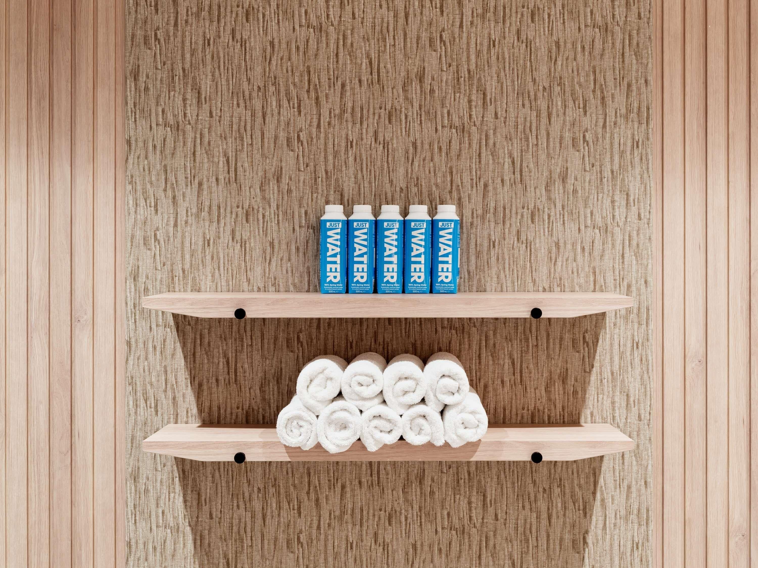 Une étagère en bois sur laquelle sont posées quatre bouteilles de « JUST WATER » et une pile de serviettes blanches.