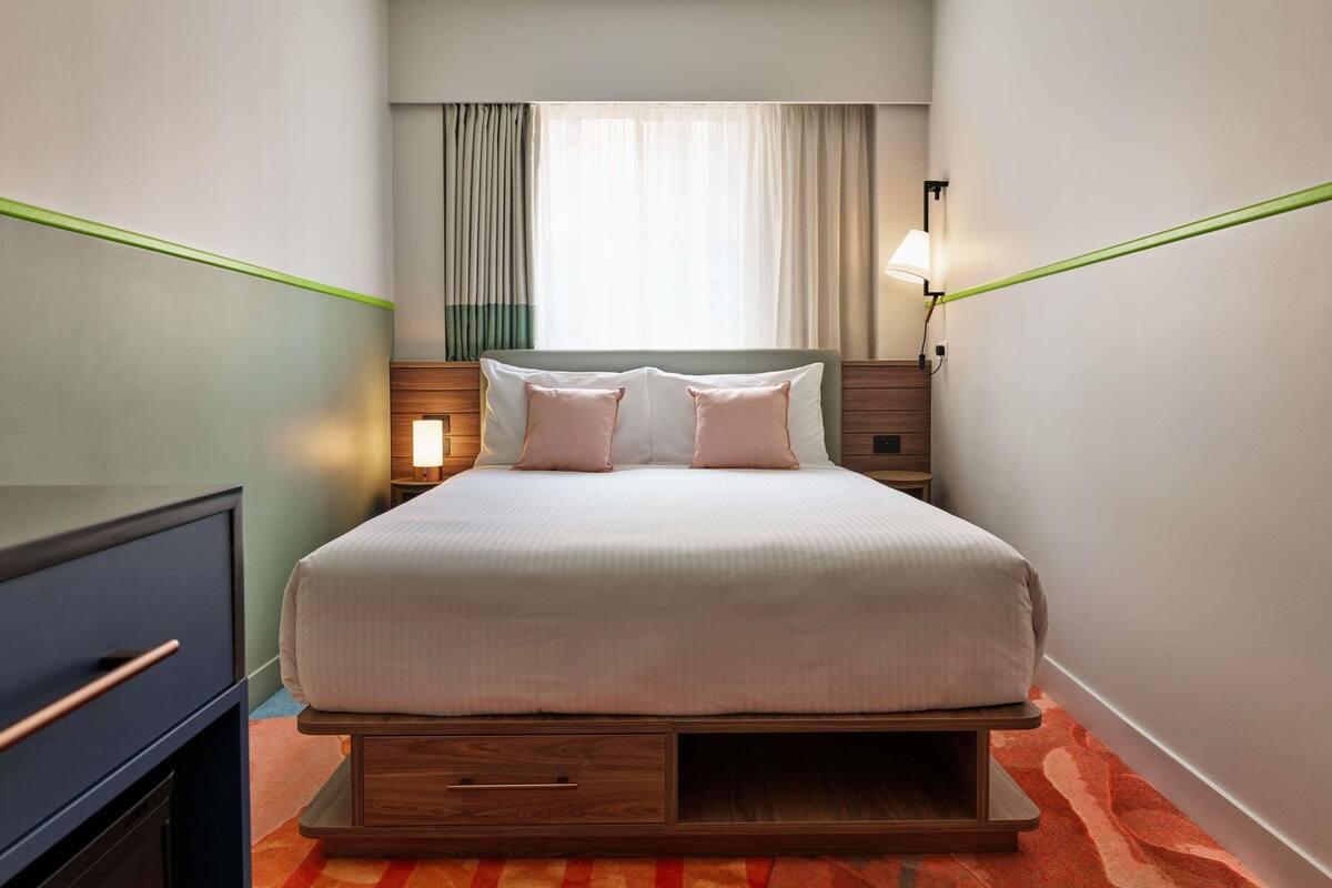 Habitación de hotel con cama, mesita de noche, lámpara y vistas al exterior a través de la ventana.