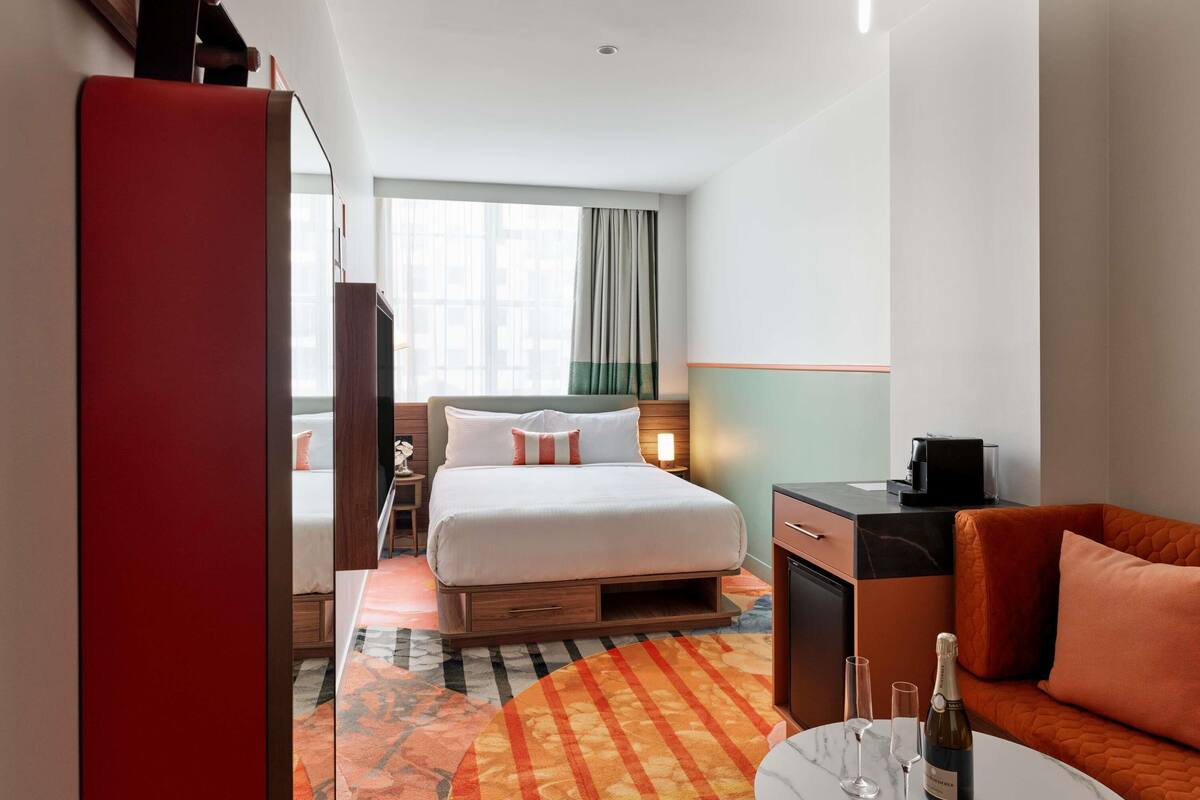 Una habitación de hotel con una cama, un escritorio, una silla, una mesita con una botella de champán y copas, y una alfombra colorida.