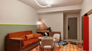 A compact room with a sofa, a small table, two chairs, a desk with a lamp, and a door.