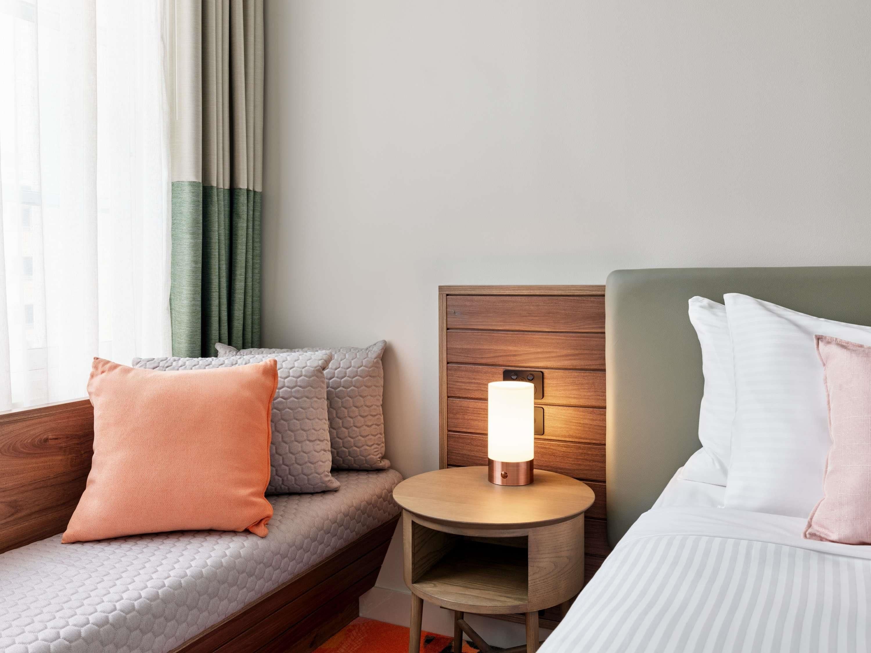 Habitación de hotel con cama, sofá, mesita de noche con lámpara y ventana con cortinas.