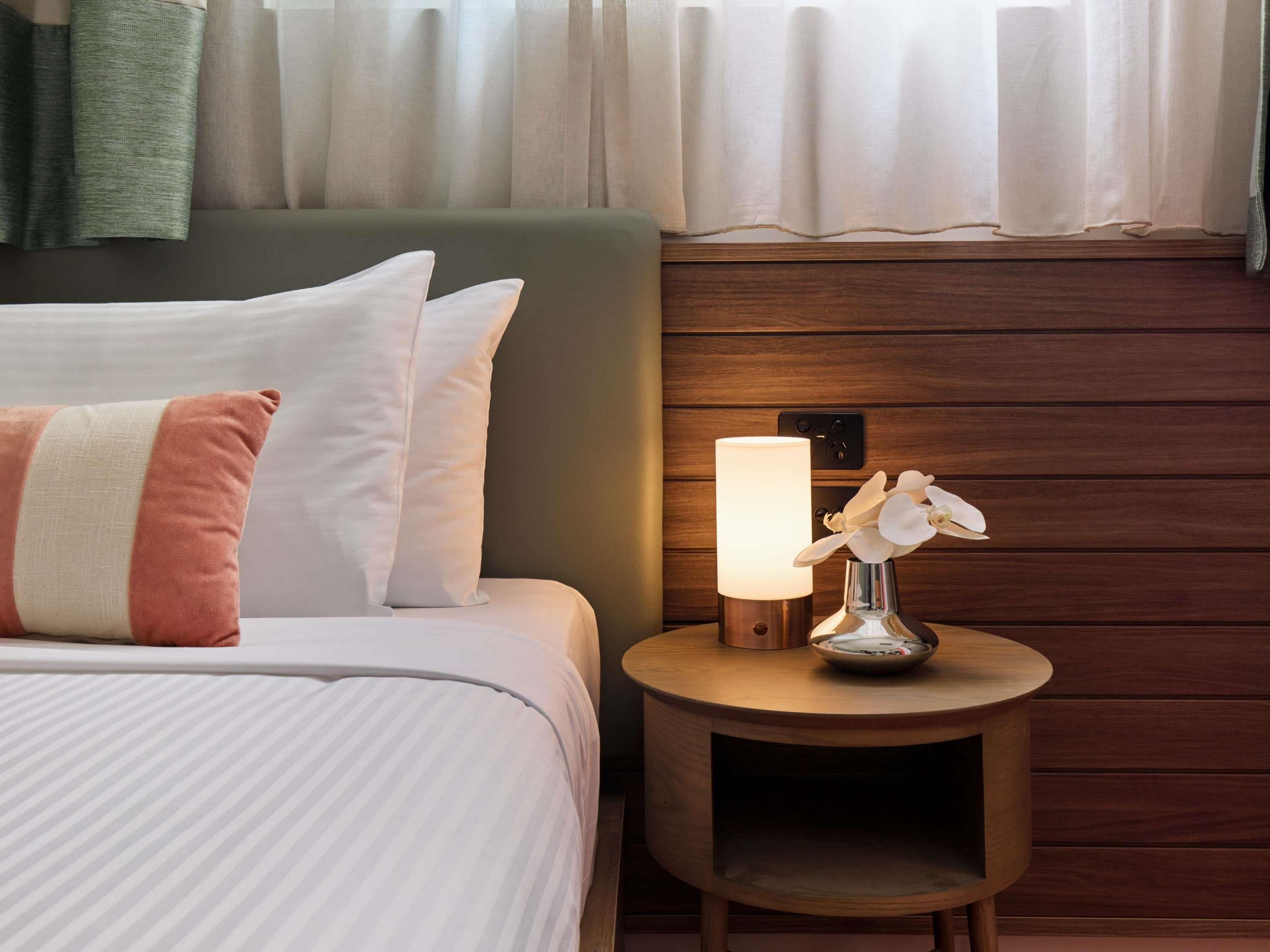 A neatly made bed with white and pink pillows, a bedside table with a lamp and orchid flowers, and wooden paneling on the wall.