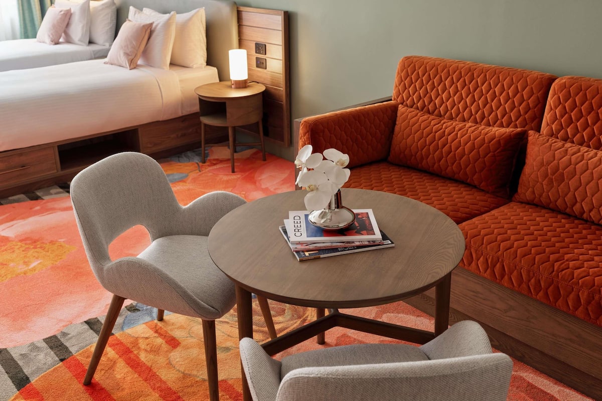 Habitación de hotel con cama, sofá, dos sillones, una mesita de noche con lámpara y un jarrón decorativo.