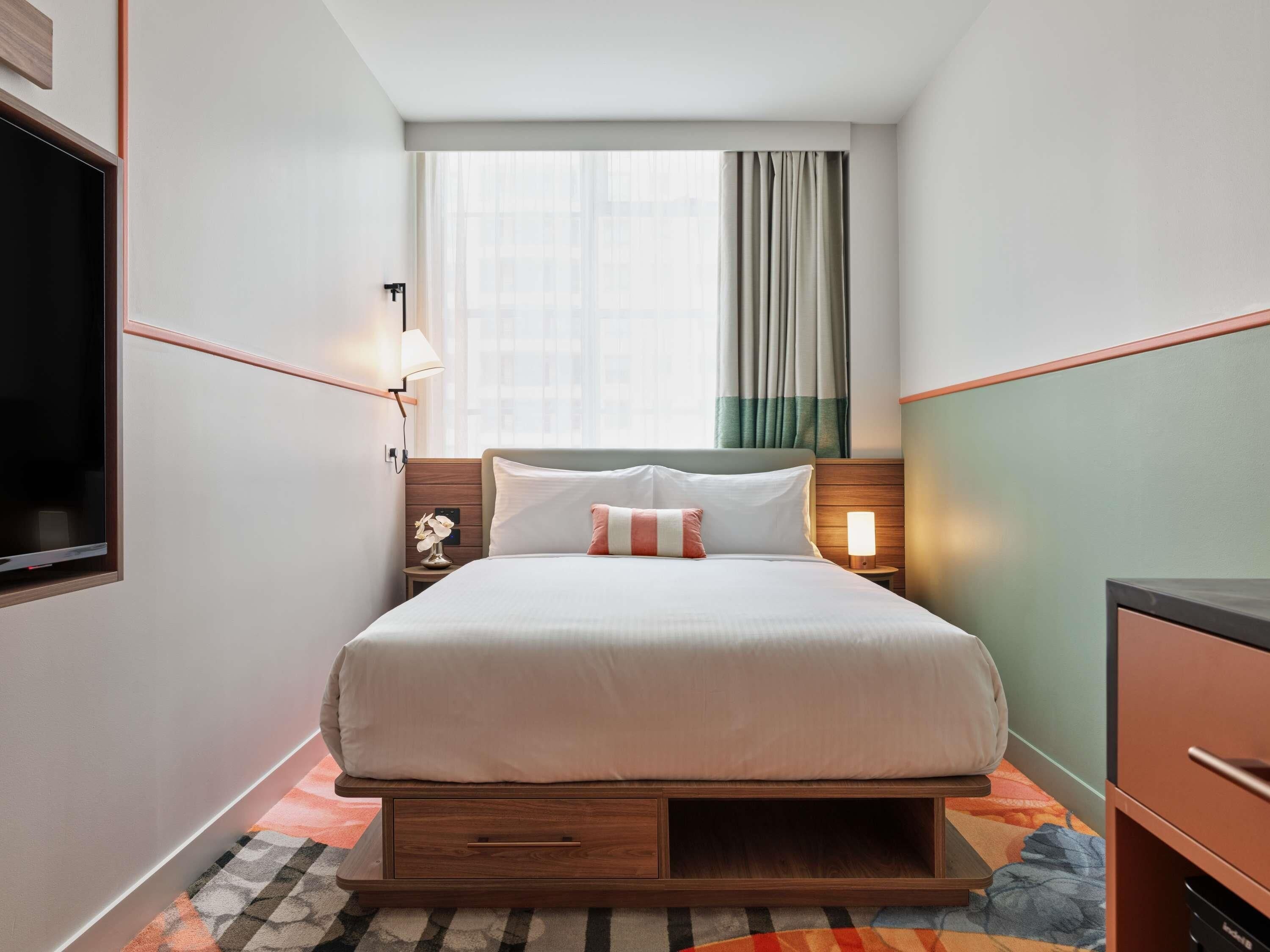 Une chambre d’hôtel de taille réduite, comprenant un lit, des tables de chevet, une télévision et une fenêtre avec des rideaux.