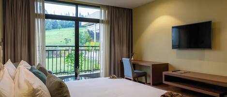 A hotel room with a large bed, a desk, a chair, a TV, and a view of a green landscape through the window.